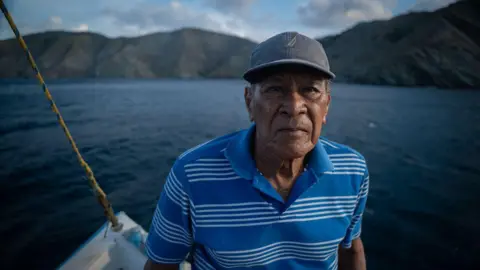 An older man with a lined face, wearing a baseball cap and a blue stripy top, is seen on board a boat with a mountainous coastline in the background.