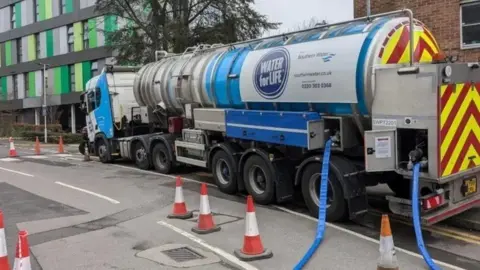 A water taker pumping out water through hoses on the street. There are orange and white road cones around it.