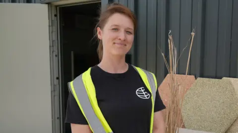 Shaun Whitmore/BBC A woman stands outside a grey building. She is smiling. She has red hair that has been tied back and is wearing a black T-shirt with a green hi-vis jacket over the top. 