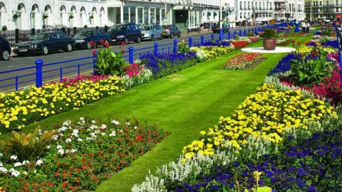 Large flowerbeds with different colours of flowers around the edge. There is a patch of green grass in the centre with a road and white buildings to the left. There is a blue metal fence between the path and flowerbed.