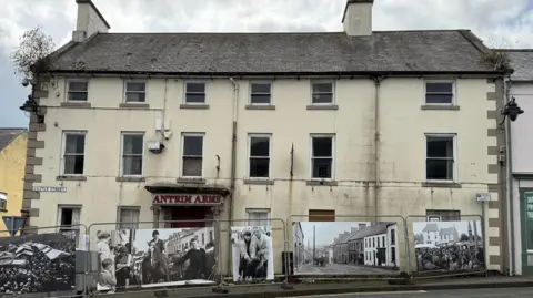The former Antrim Arms hotel in Ballycastle town centre in September 2025. The large cream-coloured building has six front windows on each of its three floors and the sign above the front door says "Antrim Arms" in red lettering. There are two chimneys and vegetation is growing out of the guttering at both sides of the roof. Two of the front windows are boarded up. Steel safety barriers, decorated with old photos of the town, stand in front of the building.