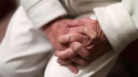 Yui Mok/PA Wire An older person dressed in a white fleecy top and white trousers sits with their hands clasped on their lap. The skin has lines and signs of sun tanning. 