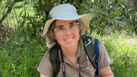 Domino Joyce A bespectacled woman with graying hair under a safari-style canvas jungle hat is crouching down in dense, lush green vegetation. She has a backpack on and a camera around her neck.
