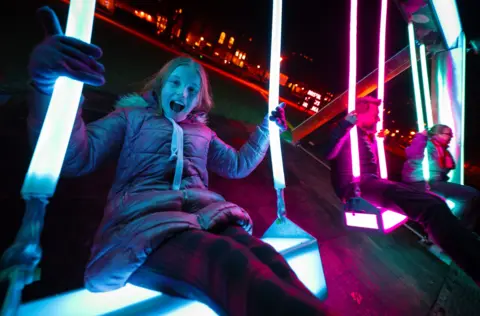 Three people swing on illuminated swings set up in Queen Square in Bristol as part of the Bristol Light Festival. The nearest one to the camera is a young woman who is smiling