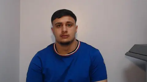 PA A young man with black hair and a thin moustache wears a blue t-shirt in a police mug shot. 