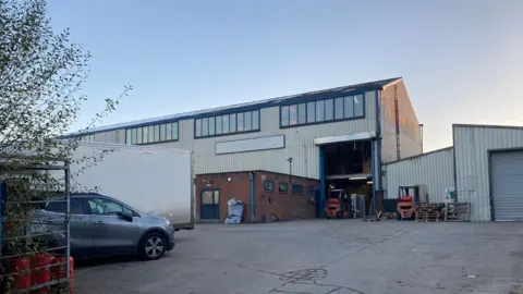 A corrugated iron warehouse building in blue and white with a large tarmac drive outside. A grey car is parked to the left