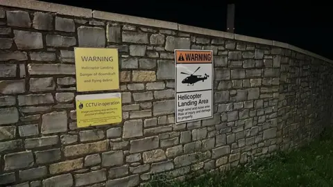 There is a sign on a brick wall which reads, 'warning, helicopter landing, danger of downdraft and flying debris' and one below which reads 'CCTV in operation' another next to it has a cartoon of a helicopter and below 'helicopter landing area, high wind and noise area, risk of injury and damage to property'