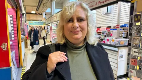 A woman with blonde hair, a green top and black jacket, with her hand on her shoulder holding her handbag, standing in Stafford indoor market.