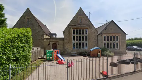 Stone school buildings with steep gabled roofs surround a fenced outdoor play area containing small plastic slides and toys.