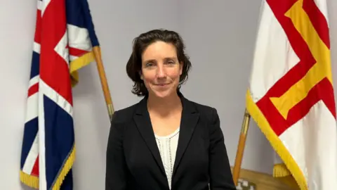 Deputy Lindsay de Sausmarez, President of the Policy & Resources Committee. A brunette woman with a black jacket on standing in front of a Union flag and a Guernsey flag.
