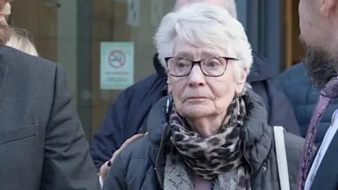 Laura Foster/BBC A grey-haired woman wearing dark-rimmed glasses and a spotted scarf and dark coat. A glass door can be seen in the background.