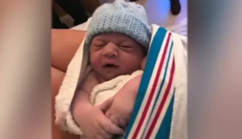 Family handout An image of a baby in a blue knitted hat wrapped in a blanket. 