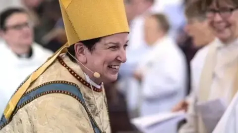 Newsline Bishop Anne Dyer dressed in full religious wear, and wearing a small head microphone, smiling while shaking hands with someone. Other people are gathered in background.
