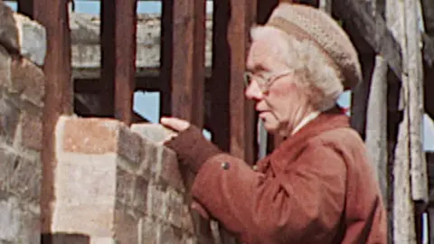 Elderly woman places a brick on a half-built house.