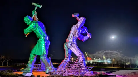 Fiona Armour The Shipbuilders of Port Glasgow sculpture - showing two men working as shipbuilders with hammers raised in the air - lit up by a bright moon