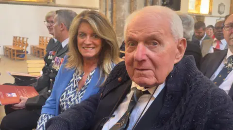 A man with white hair, wearing a dark tie and a woman with long blonde hair wearing a blue top