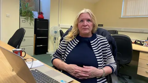 Christine Nicholls is sitting at a desk with a laptop in front of her. She is wearing a black and white striped top and a blue jumper.