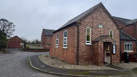 Halls A brick church with a porch at the front and windows, with a road winding round it to the left and a house and garage behind.