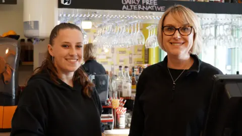 Christina and Claire are both wearing black tops and standing behind the coffee shop counter, smiling. In the background is a mirror and hanging up wine glasses.