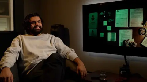 A man with a beard wearing a grey sweatshirt and dark trousers sitting in a chair next to a noticeboard