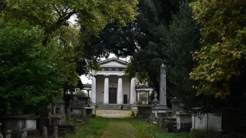 Getty Images A tree-lined path leads to a grand white building with tall columns and a triangular pediment. Surrounding the path are numerous old stone tombs and monuments in Kensal Green cemetery. 