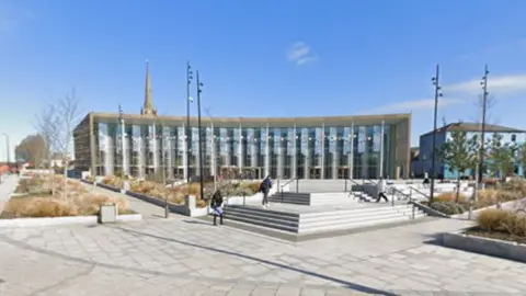 Google A university building with a glass front. Students are walking up steps on the public square in front of it. There are a few edged-off planted sections with dry, scrubby grass and plants in them. The sky is a brilliant blue. 