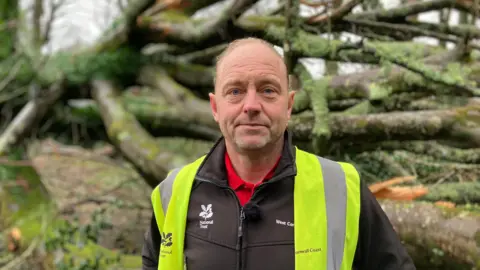 A photo of Simon Hocking - he is standing in front of a tree that has been blown over. He is wearing a high-viz jacket and black waterproof coat. The logo on his coast says National Trust.