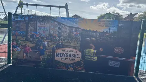 Banner on a play park fence, showing children playing sunshine and men being rejected at a barrier. 