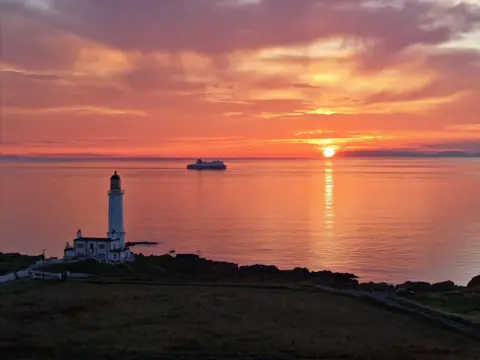Rob Barrie A vivid orange and yellow sun setting in the distance with a lighthouse in the foreground