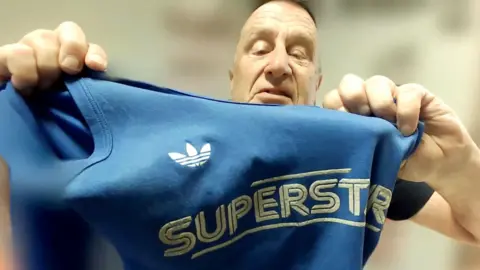 Mark Ashwell holds a blue shirt with the word "Superstars" close to the camera.
