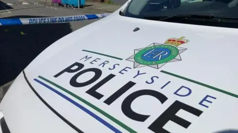 A close up of the front of a Merseyside Police vehicle with the force's badge and the words Merseyside Police displayed prominently on the bonnet in bold blue and black font. In the background there is blue tape which reads 'police line, do not cross'.