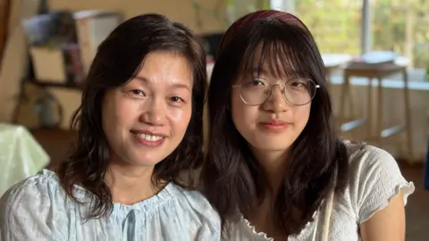 Rita and Sheren smile at the camera. Sheren, on the right, is wearing round glasses. They both have dark hair and are wearing light coloured shirts. 