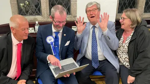 BBC Four people - three men and a woman - wearing smart clothing sit on chairs to look at a laptop. One of the men is holding his hands up.