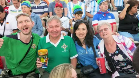 Jonny Blair A crowd at a football match, four are looking at the camera, two men are wearing green tops and one women is wearing a blue jacket and another is wearing a pink white and purple jacket with black floral top