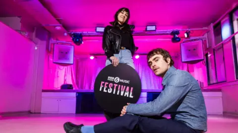 Two people holding a sign that reads: BBC Radio 6 Music Festival. The lighting is pink and this is a posed shoot.