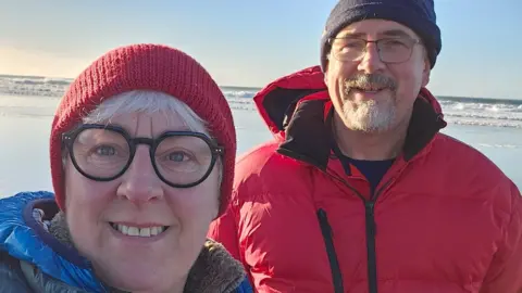 University of Aberdeen Dogwalkers Jenny Snedden and Ivor Campbell pictured on a sunlit beach wearing winter hats and jackets