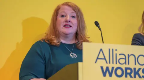 PA Media Naomi Long delivering a speech behind a yellow Alliance Works plinth