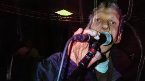 Ben G. Brown An older man with grey hair holds a microphone on a stand close to his face. He wears a black jacket, brown jumper and white t-shirt. Behind him, stage rigging and an overhead light can be faintly seen. 