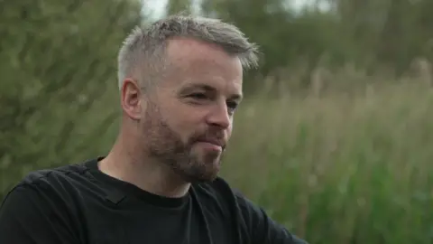 A man with light hair and a dark beard stands in front of piece of green land.