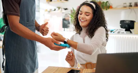 A curly brown haired woman uses her watch for card payment in a cafe - she has a laptop in front of her. her phone lies on the table. She wears a white top, brown belt and white headband. 