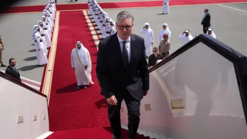 Keir Starmer boarding a plane at the airport in Doha, Qatar. There is a red carpet, lined with men, leading up to the plane