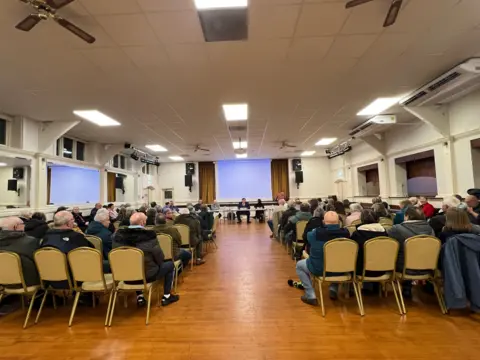 Around 100 people seated in a room with wooden flooring and a panel of councillors at the front infront of a stage 