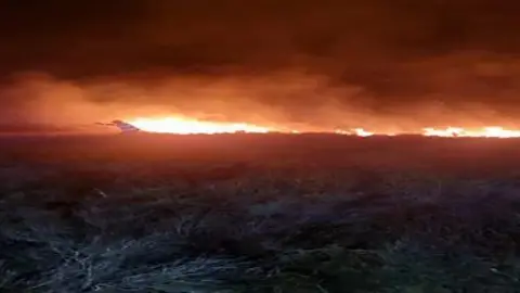 A large fire against the backdrop of a dark night sky. It is on a hill with gorse and grass spreading out in front of it.