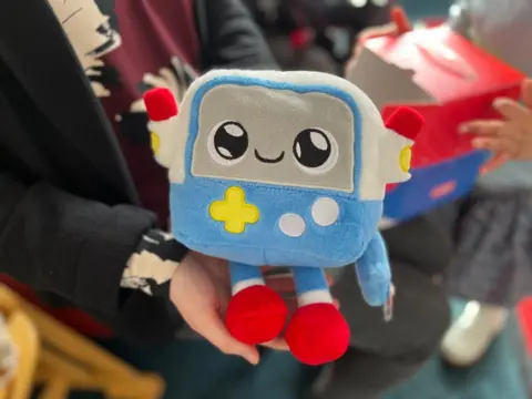 Gabbo, a cuddly toy, is held in somebody's hands. Gabbo is soft-textured, like a teddy bear, but looks like a robot.