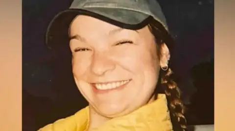 Clare Dupree, smiling, and wearing a yellow coat and green baseball cap. She has brown plaited hair. 
