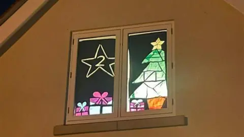 Resident Kate A house window with a decoration of a Christmas tree and presents underneath. The number 2 is displayed and surrounded with a star.