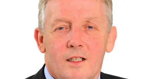 Tameside Council Close up of Ged Cooney with grey hair and blue eyes wearing a pale blue shirt and black suit jacket with a white background