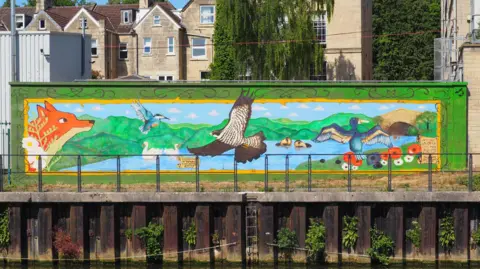 Colourful mural of animals and a river, painted on the side of a building, with houses behind it. The mural shows a fox, a duck, swans, fish, a kingfisher, otters and a bird of prey. It includes a river with a bridge over it that joins two sides of grassland.