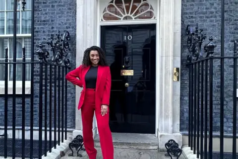 Jamilah Hassan Jamilah Hassan is pictured standing outside No.10 Downing Street. She is wearing a red trouser suit and has black coiled hair. 