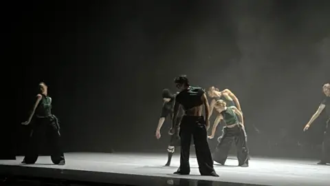 BBC Six dancers on a stage. They are wearing green tops and black bottoms and are learning towards stage right. 